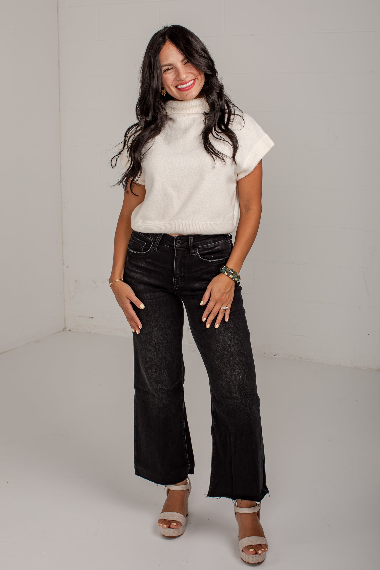 A woman modeling a white short-sleeve mock neck knit top and black wide-leg cropped jeans with white heeled sandals, standing against a plain light gray studio background.
