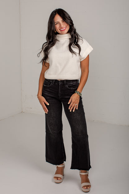 A woman modeling a white short-sleeve mock neck knit top and black wide-leg cropped jeans with white heeled sandals, standing against a plain light gray studio background.