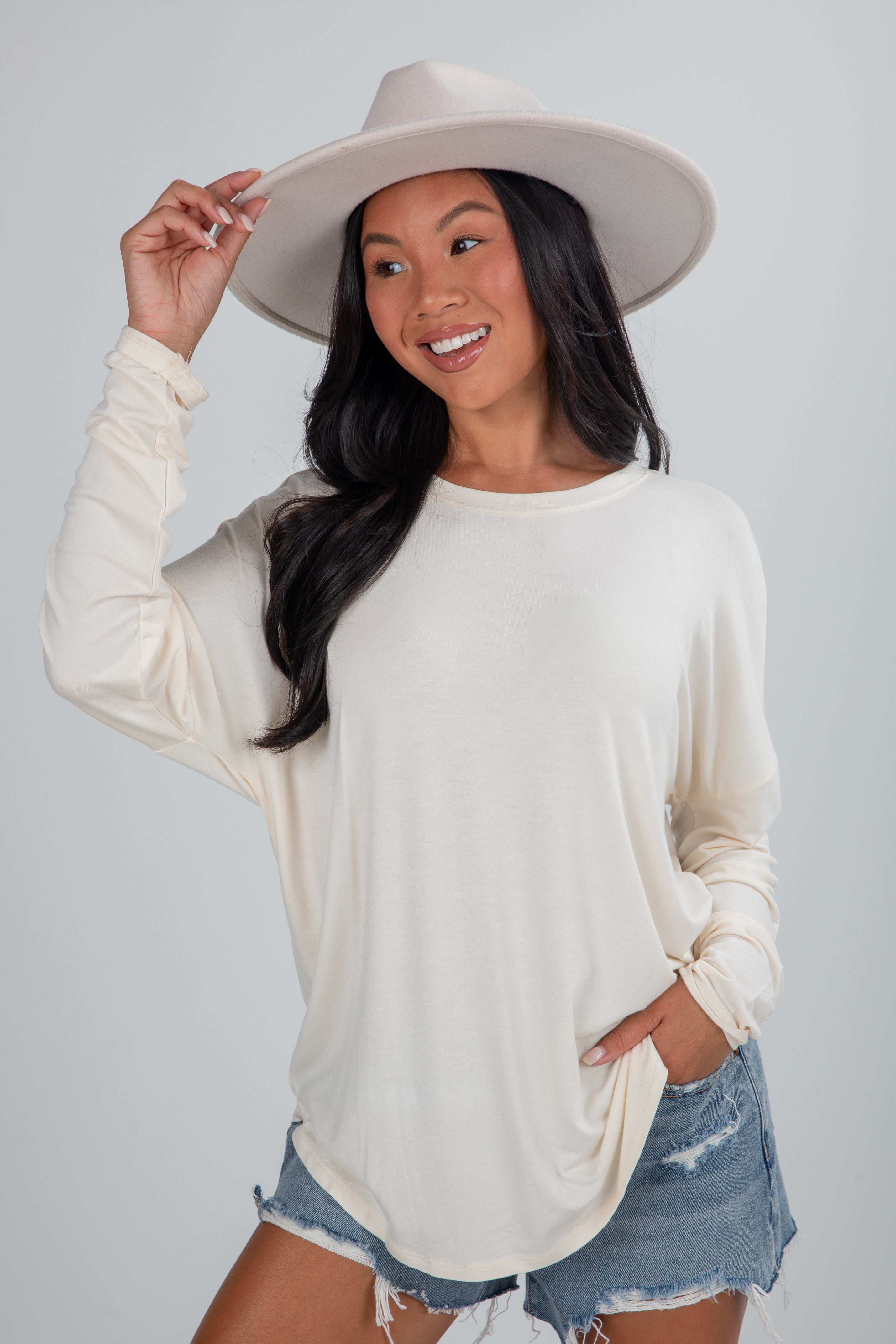 Woman wearing a white long-sleeve top and denim shorts with a white hat on a plain background
