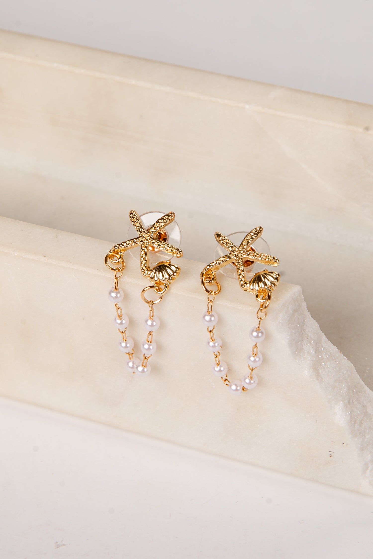 Gold earrings with starfish designs and pearl strands on a beige surface