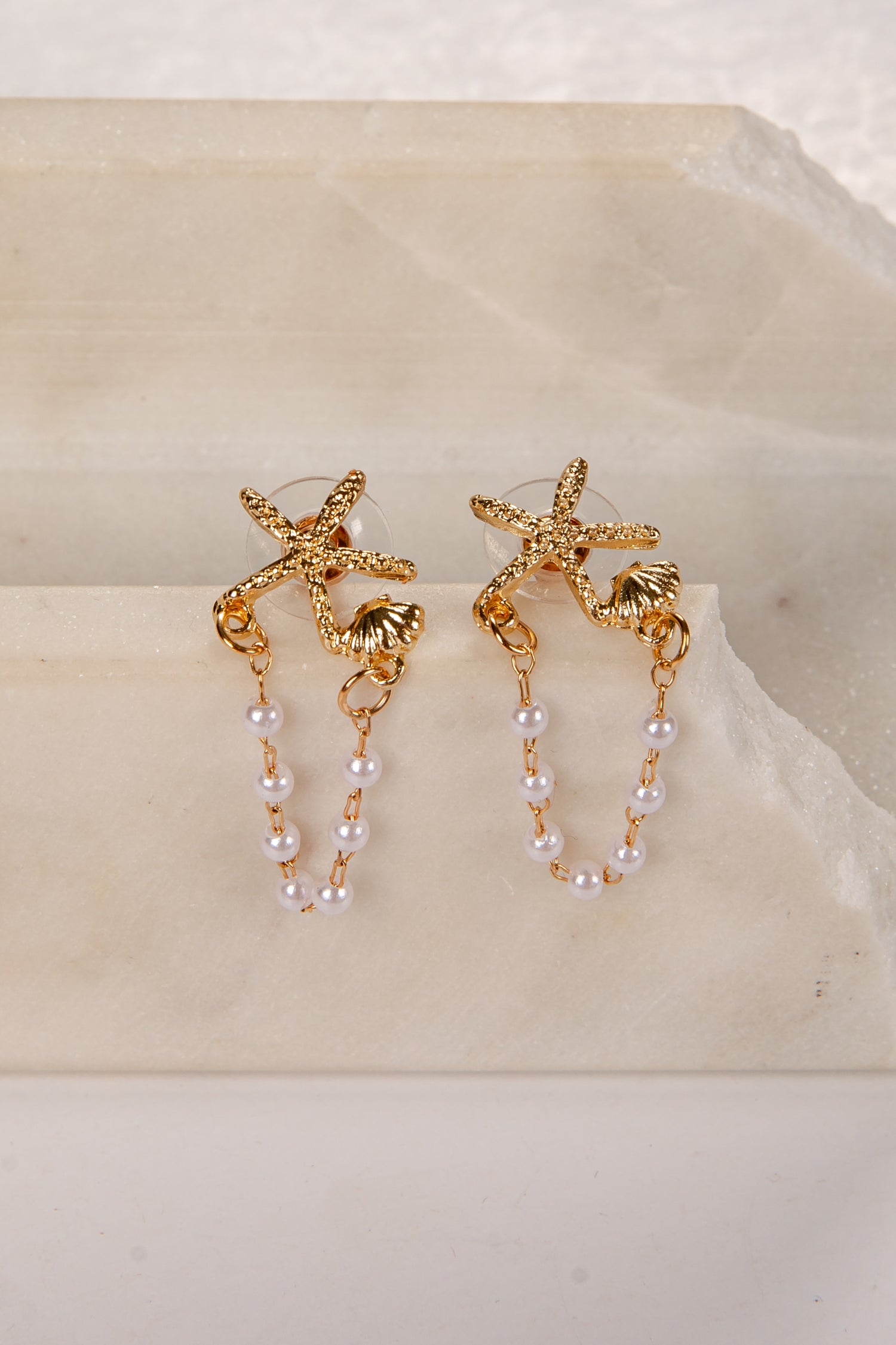 Gold starfish earrings with pearl chains on a beige stone surface