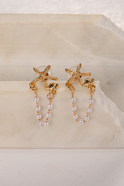 Gold starfish earrings with pearl chains on a beige stone surface