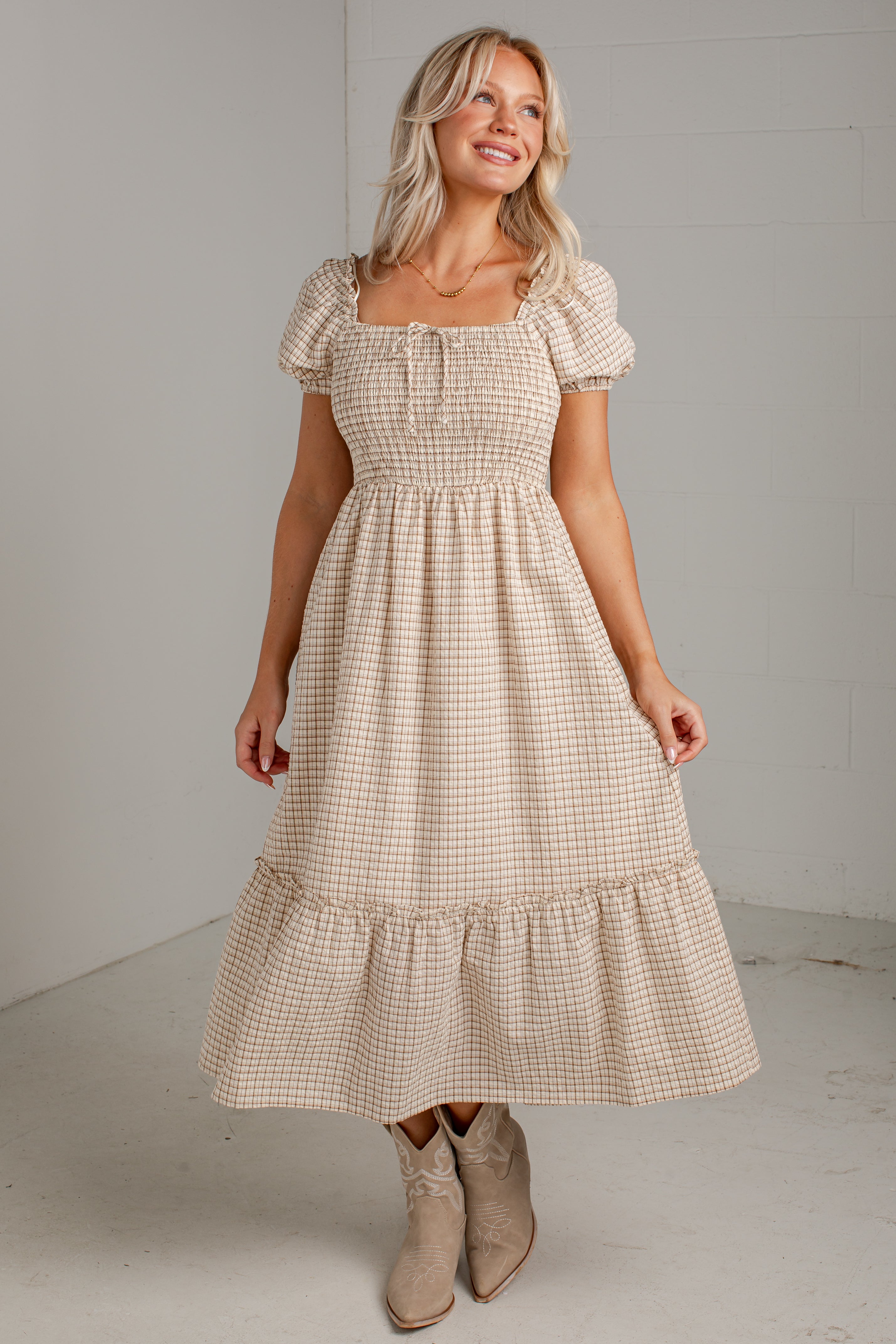 A woman modeling a beige and white gingham midi dress with puff sleeves, a smocked bodice, and a tiered skirt, paired with beige cowboy boots in a studio setting.