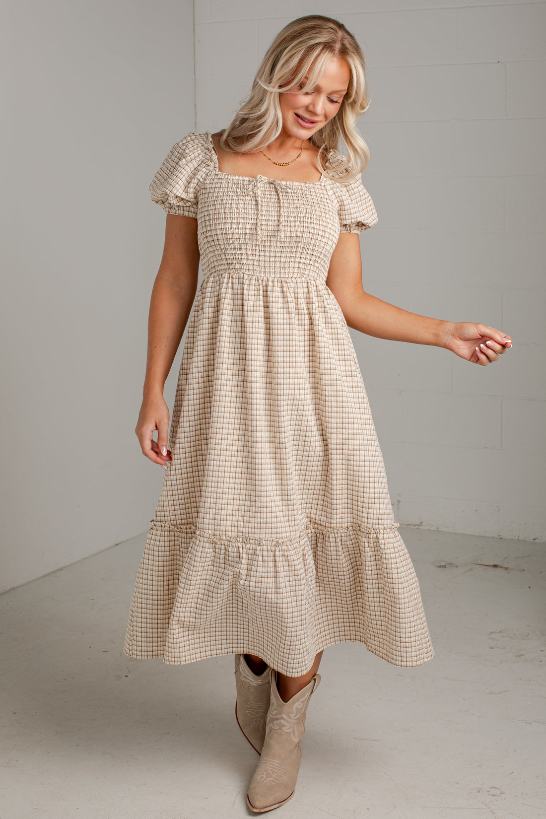 A woman modeling a beige and white gingham tiered midi dress with puff sleeves and a smocked bodice, paired with beige ankle boots, in a minimalist studio setting.
