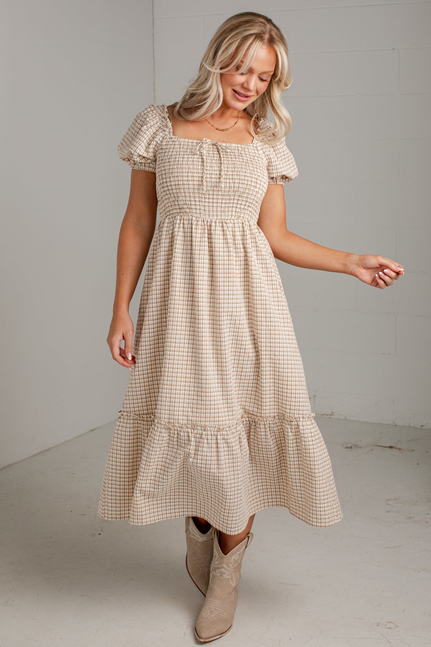 A woman modeling a beige and white gingham tiered midi dress with puff sleeves and a smocked bodice, paired with beige ankle boots, in a minimalist studio setting.
