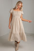 A woman modeling a beige and white gingham tiered midi dress with puff sleeves and a smocked bodice, paired with beige ankle boots, in a minimalist studio setting.