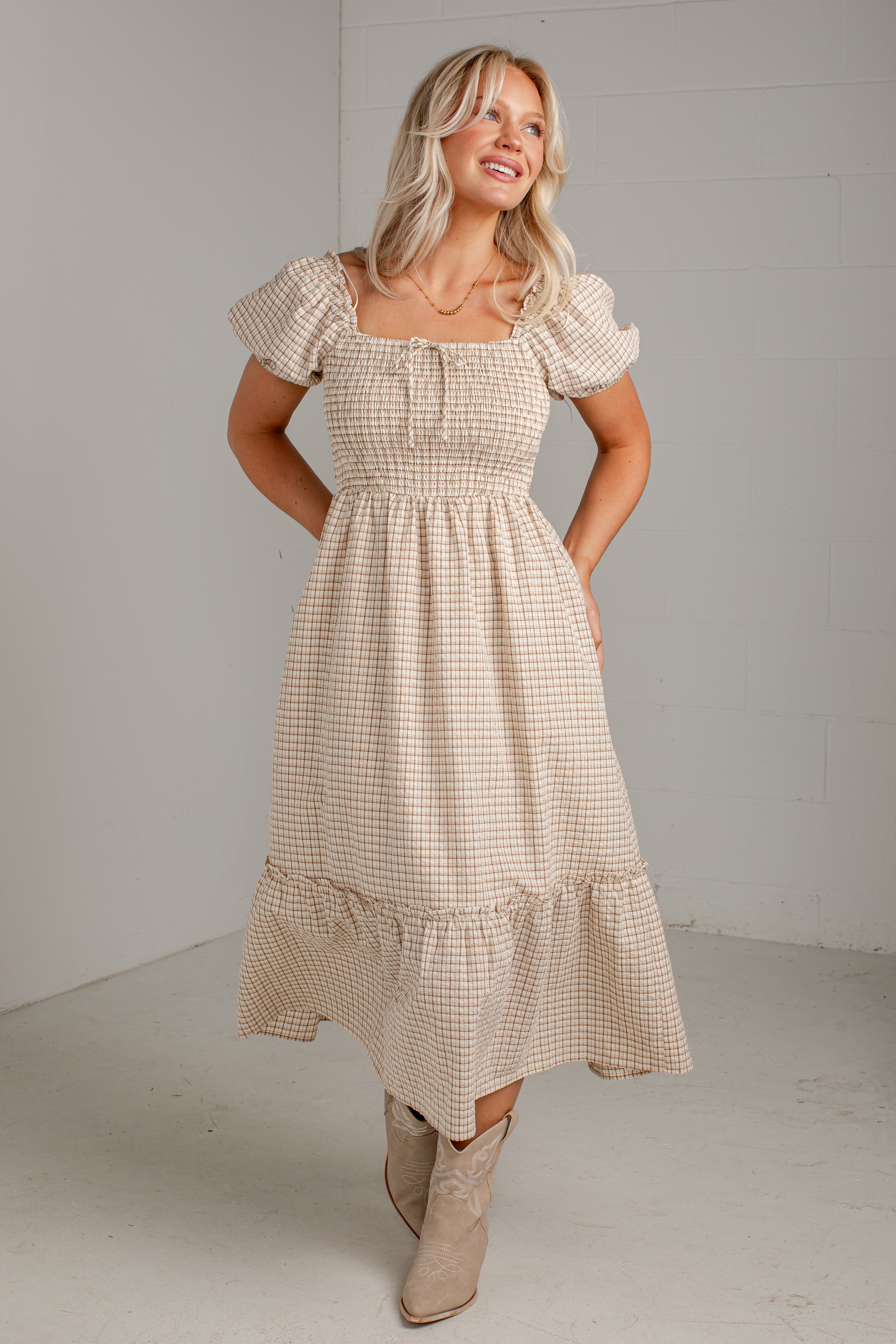 A woman modeling a beige and white gingham tiered midi dress with puff sleeves and a smocked bodice, paired with beige ankle boots, in a minimalist studio setting.