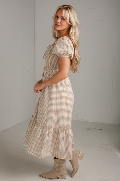 A woman modeling a beige and white gingham tiered midi dress with puff sleeves and a smocked bodice, paired with beige ankle boots, in a minimalist studio setting.