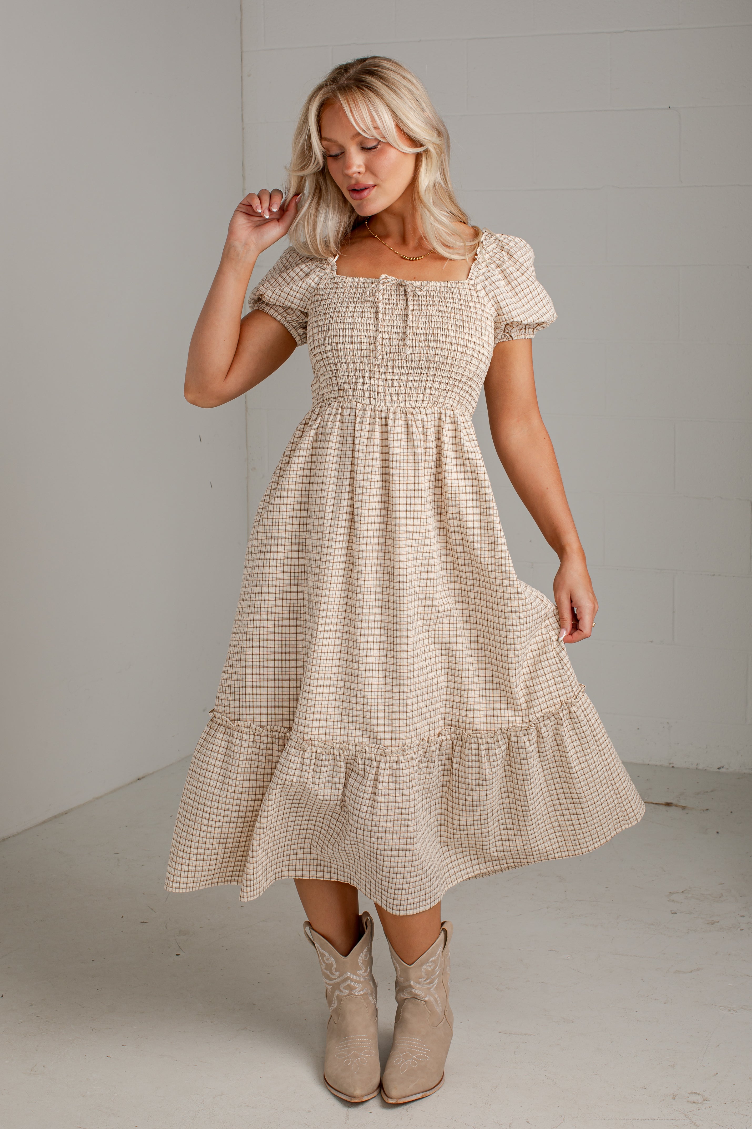 A woman modeling a beige and white gingham tiered midi dress with puff sleeves and a smocked bodice, paired with beige ankle boots, in a minimalist studio setting.