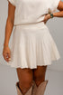 A model wearing a cream-colored pleated knit mini skirt with a matching knit top and tan cowboy boots, shown from the chest down against a plain light gray background.