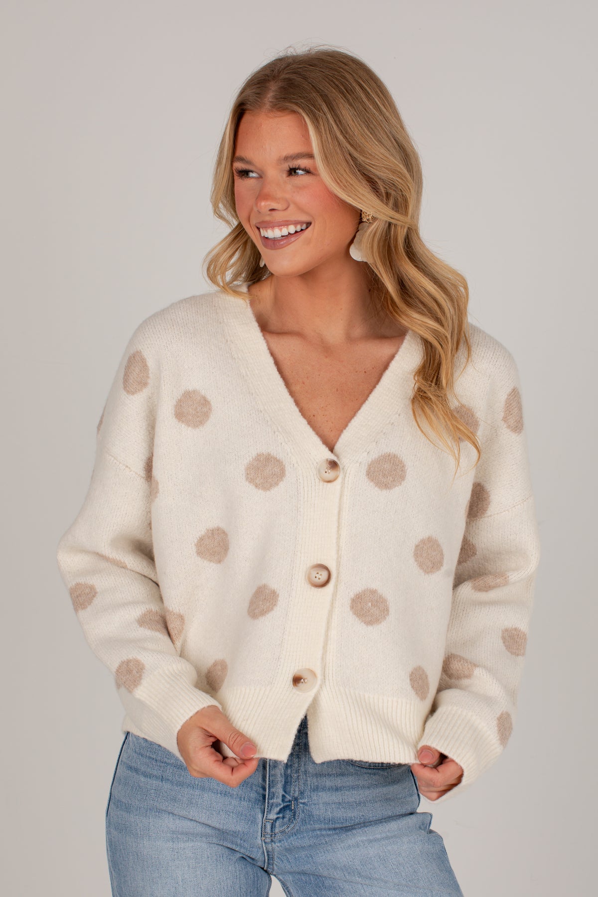 A smiling blonde woman wearing a cream-colored knit cardigan with beige polka dots and light blue jeans, photographed against a solid light gray studio background.