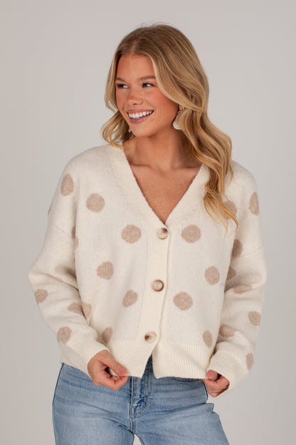 A smiling blonde woman wearing a cream-colored knit cardigan with beige polka dots and light blue jeans, photographed against a solid light gray studio background.