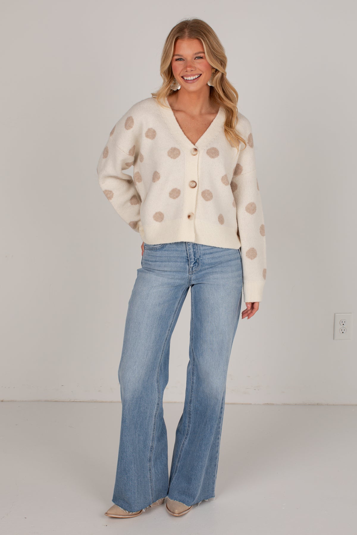 A woman modeling a cream-colored cardigan sweater with beige polka dots and light-wash blue flare jeans, standing against a plain light gray wall.