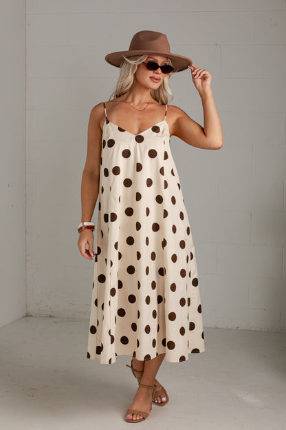 A woman modeling a cream-colored midi slip dress with dark brown polka dots, styled with a brown wide-brim hat, sunglasses, and brown strappy heels against a light gray studio background.