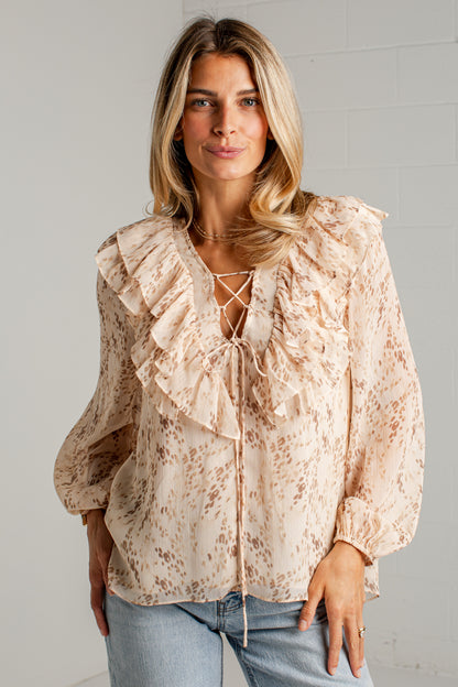 A woman wearing a beige and brown leopard-print blouse with layered ruffles and lace-up front details, paired with light blue jeans, standing against a white brick wall.