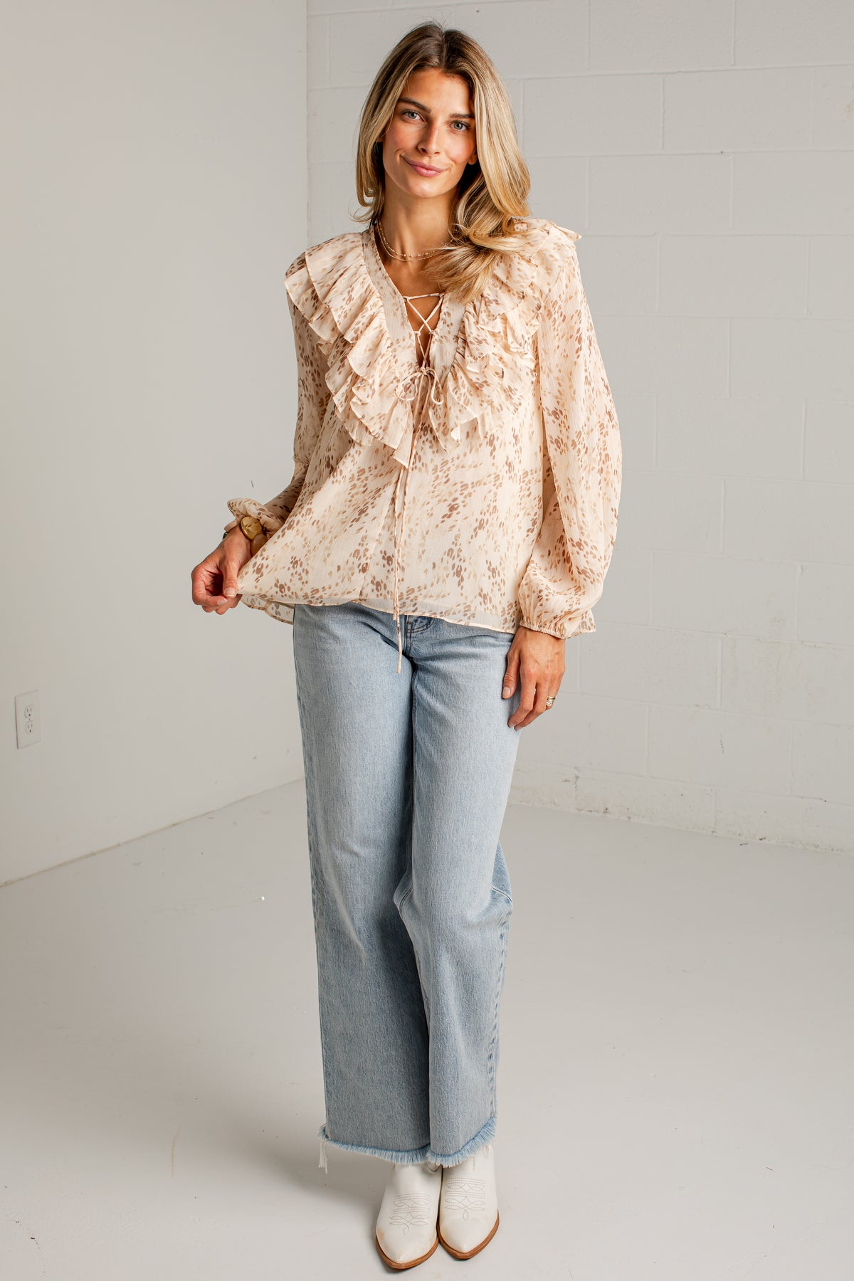 A woman modeling a light beige blouse with ruffles and a tie neckline, paired with light blue wide-leg jeans and white ankle boots, standing in a bright white studio.