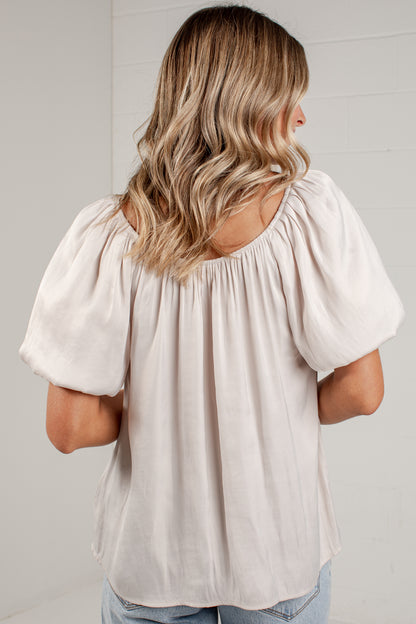A woman viewed from behind wearing a light beige puff-sleeve top and light blue jeans, standing against a white paneled wall.
