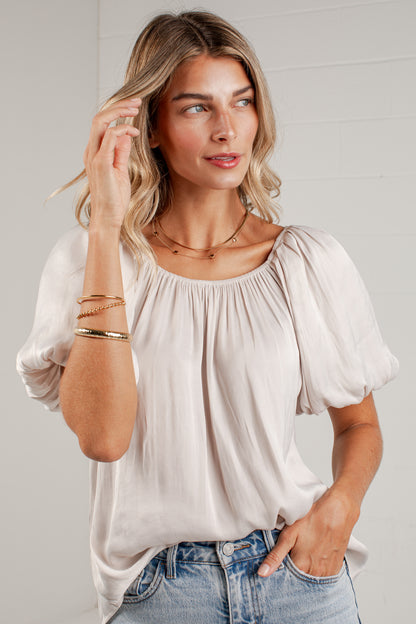A woman wearing a light beige puff-sleeve blouse with a gathered neckline, paired with blue jeans, standing against a light gray brick wall.