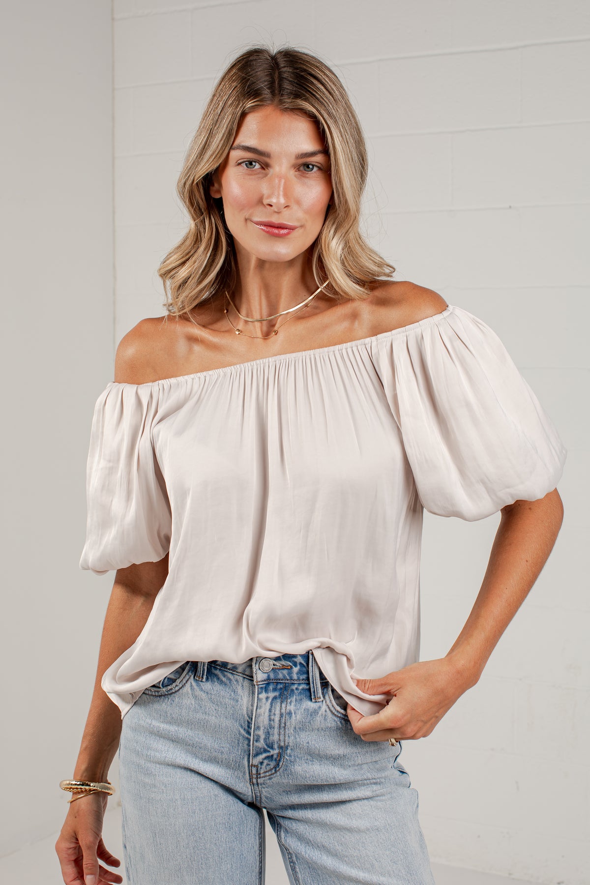 A woman modeling an off-the-shoulder cream puff-sleeve blouse with light-wash blue jeans, standing against a white brick wall.