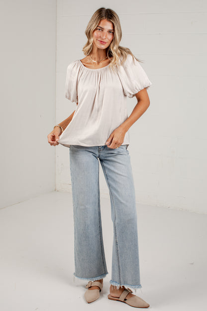 A woman modeling a light beige short-sleeve blouse with puff sleeves and light-wash wide-leg jeans, standing against a white studio wall.