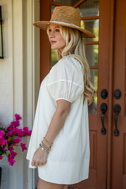 A woman wearing a cream puff-sleeve tunic dress and a straw hat, standing in profile in front of a brown wooden door with pink flowers on the left.