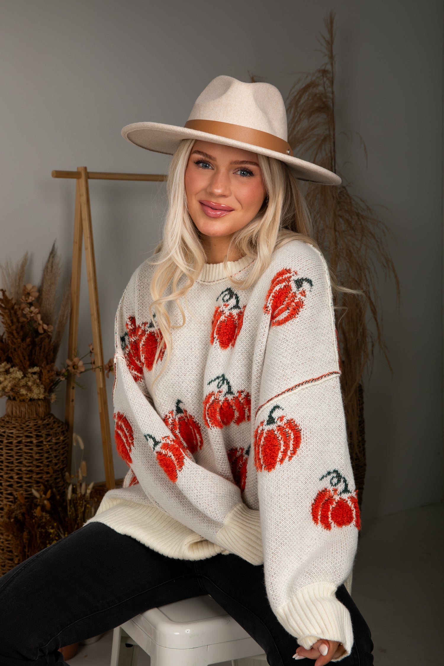 Woman wearing a white sweater with red pumpkin patterns and a beige hat, sitting on a stool.
