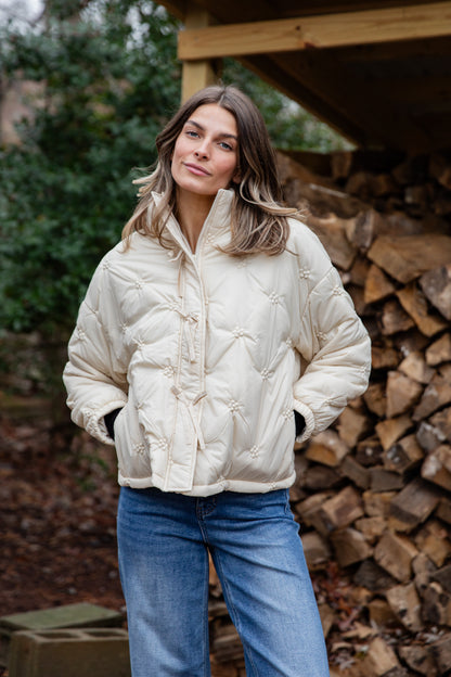 Woman wearing a cream puffer jacket and blue jeans standing in front of stacked firewood.