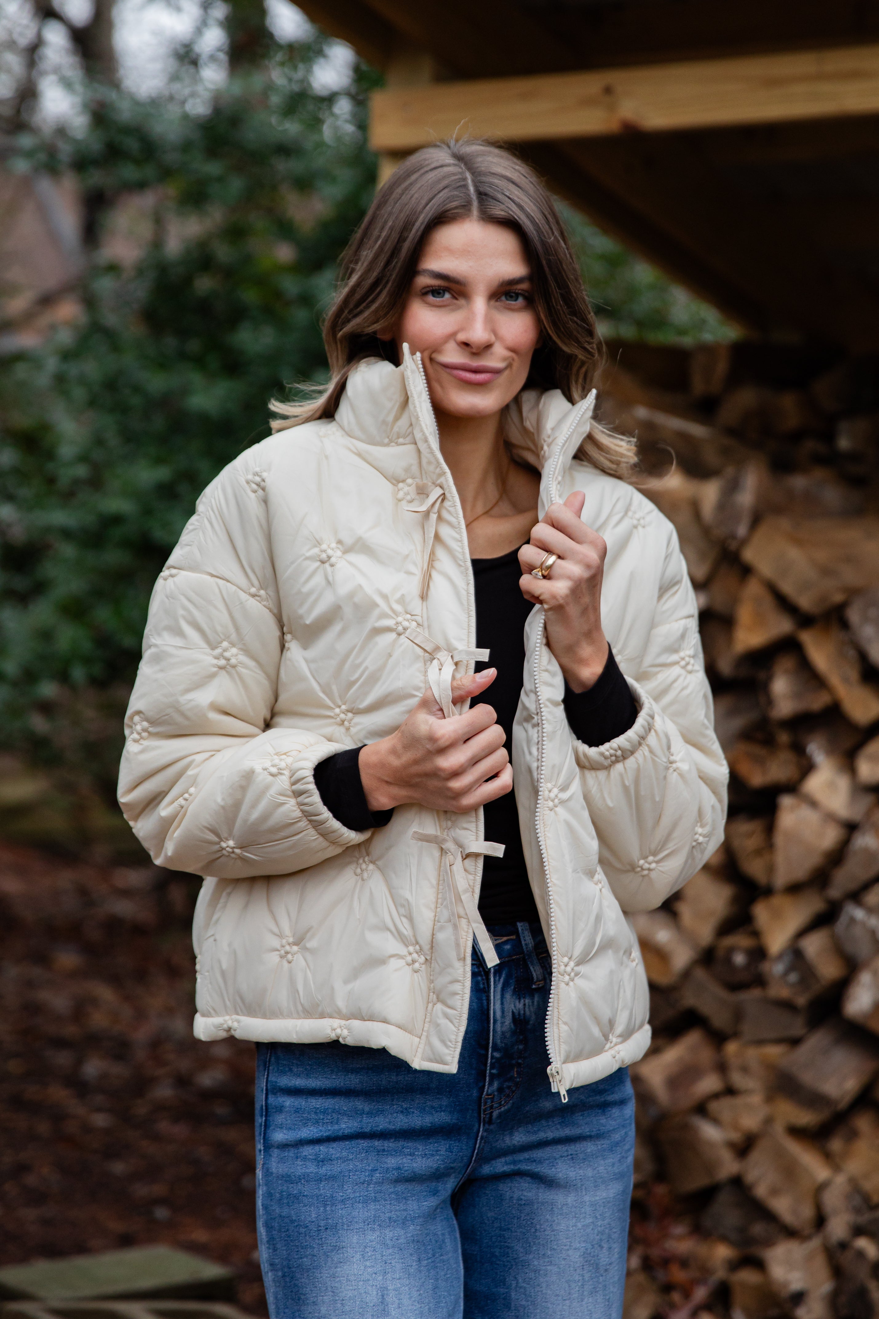 Woman wearing a cream puffer jacket standing in front of stacked firewood.