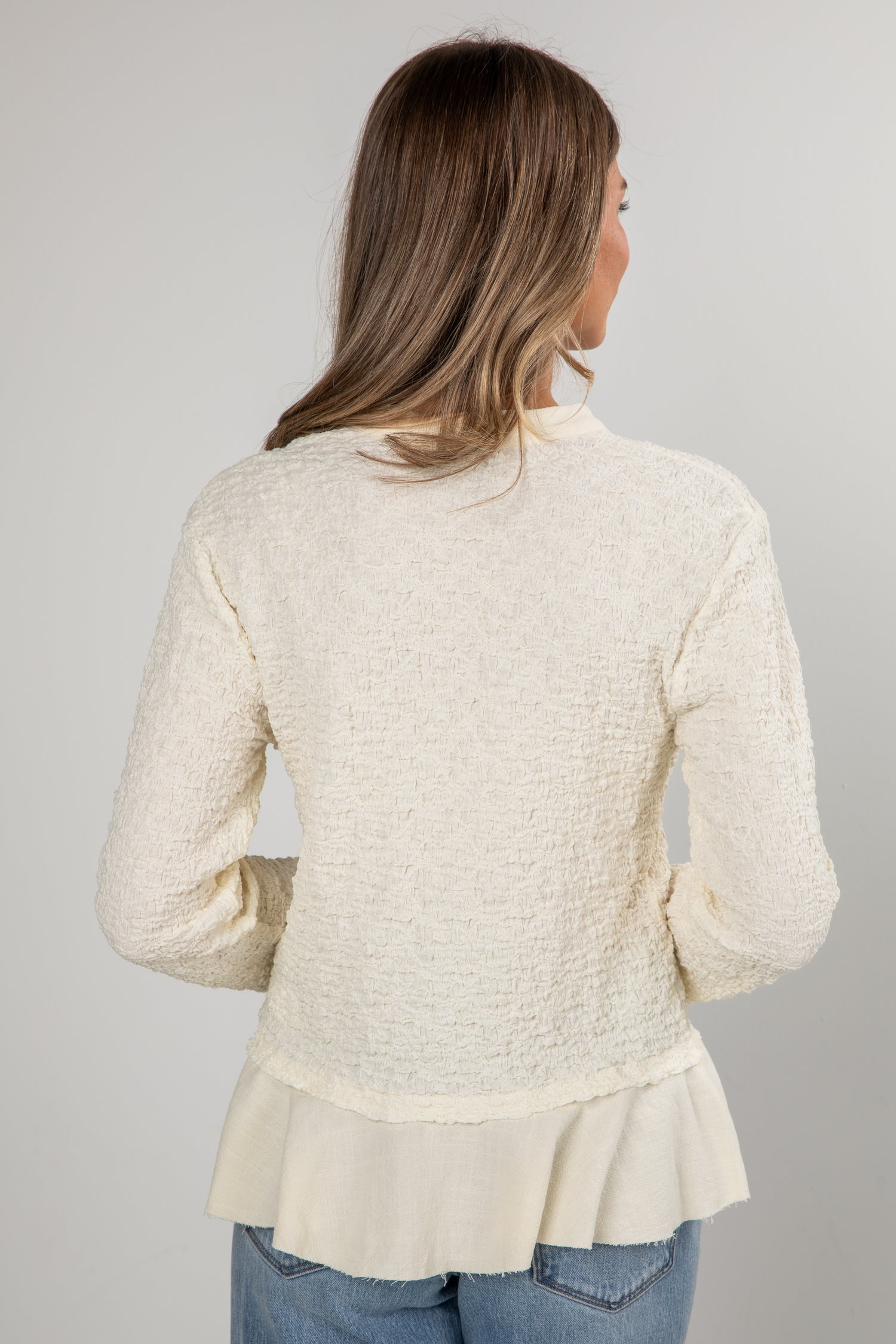 Woman wearing a textured beige cardigan with a ruffled bottom on a gray background