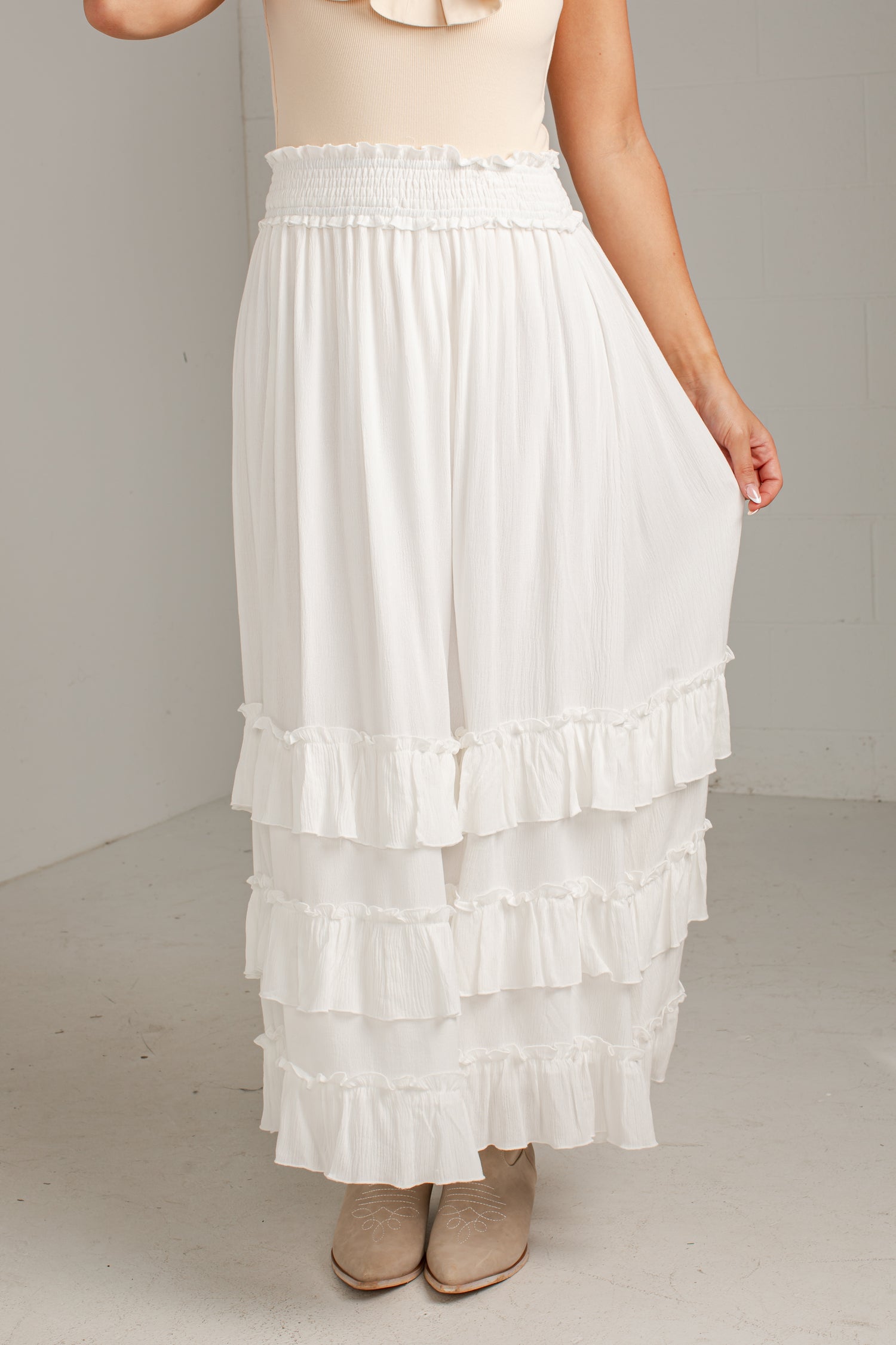 A white tiered maxi skirt with a smocked elastic waistband, shown on a model wearing beige ankle boots against a neutral studio background.