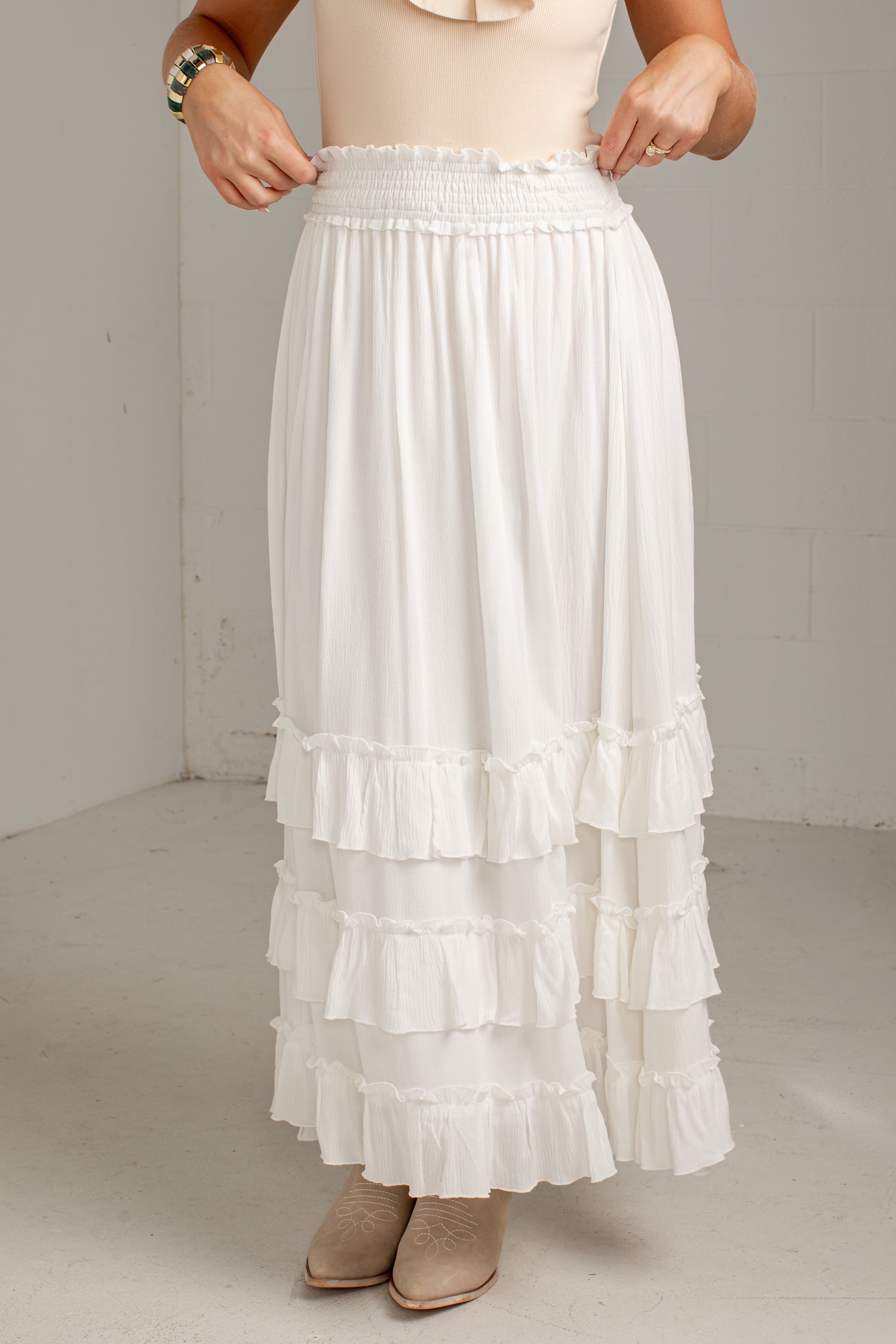 A white tiered maxi skirt with a smocked elastic waistband, shown on a model wearing beige boots and a cream top in a neutral studio setting.