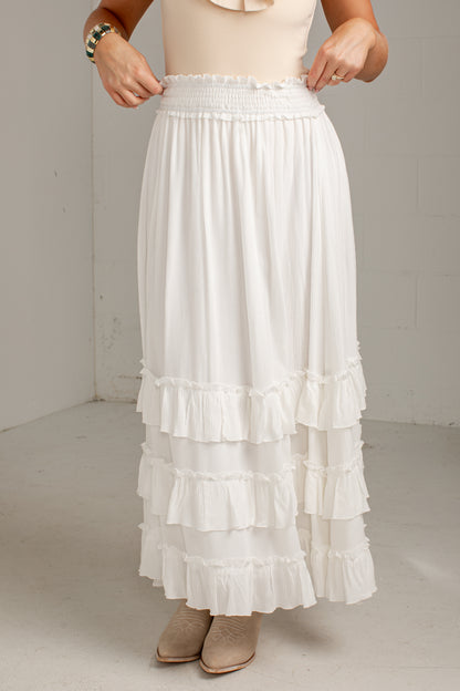 A white tiered maxi skirt with a smocked elastic waistband, shown on a model wearing beige boots and a cream top in a neutral studio setting.