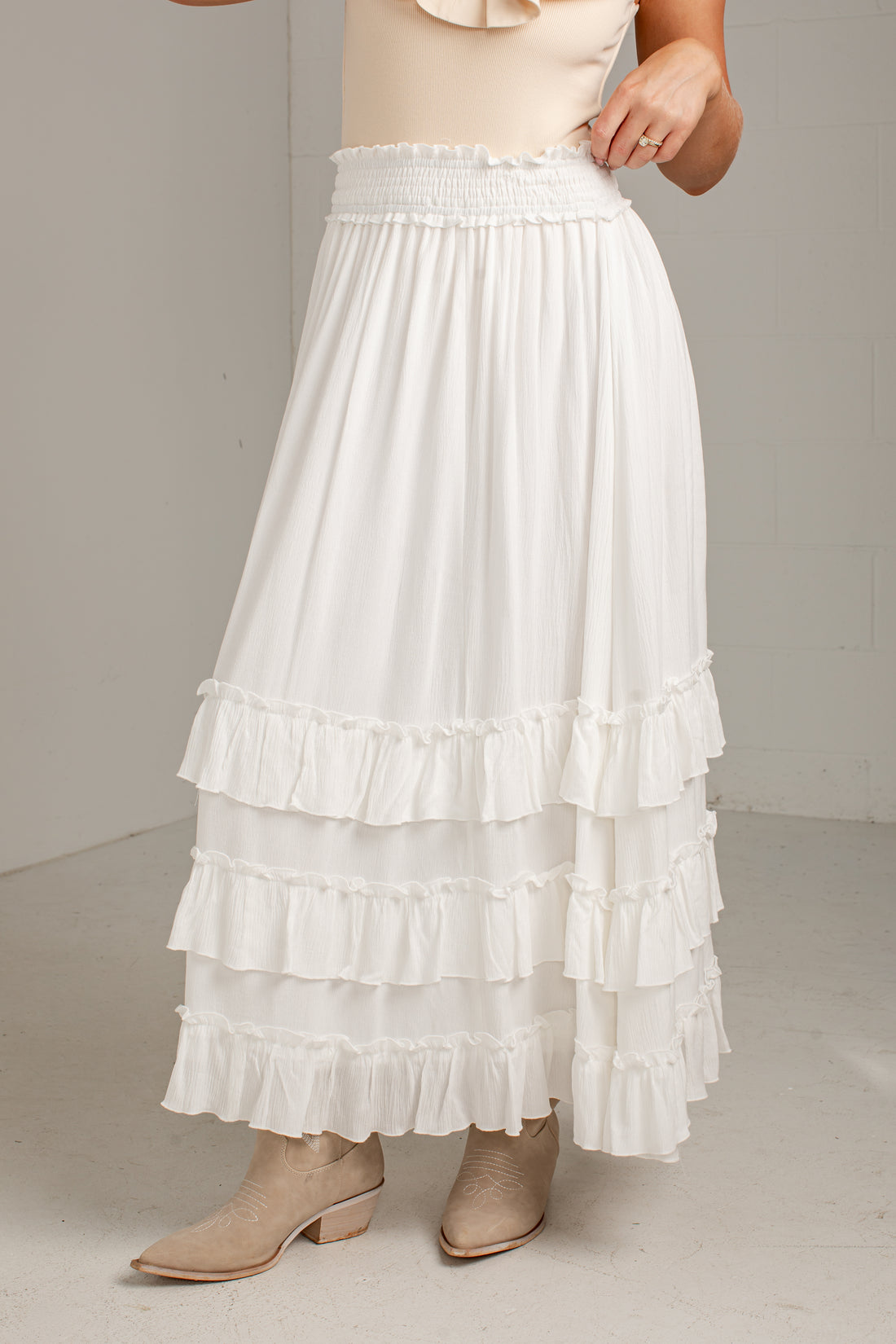 A white tiered maxi skirt with an elastic smocked waistband, shown on a model wearing beige ankle boots against a neutral studio background.