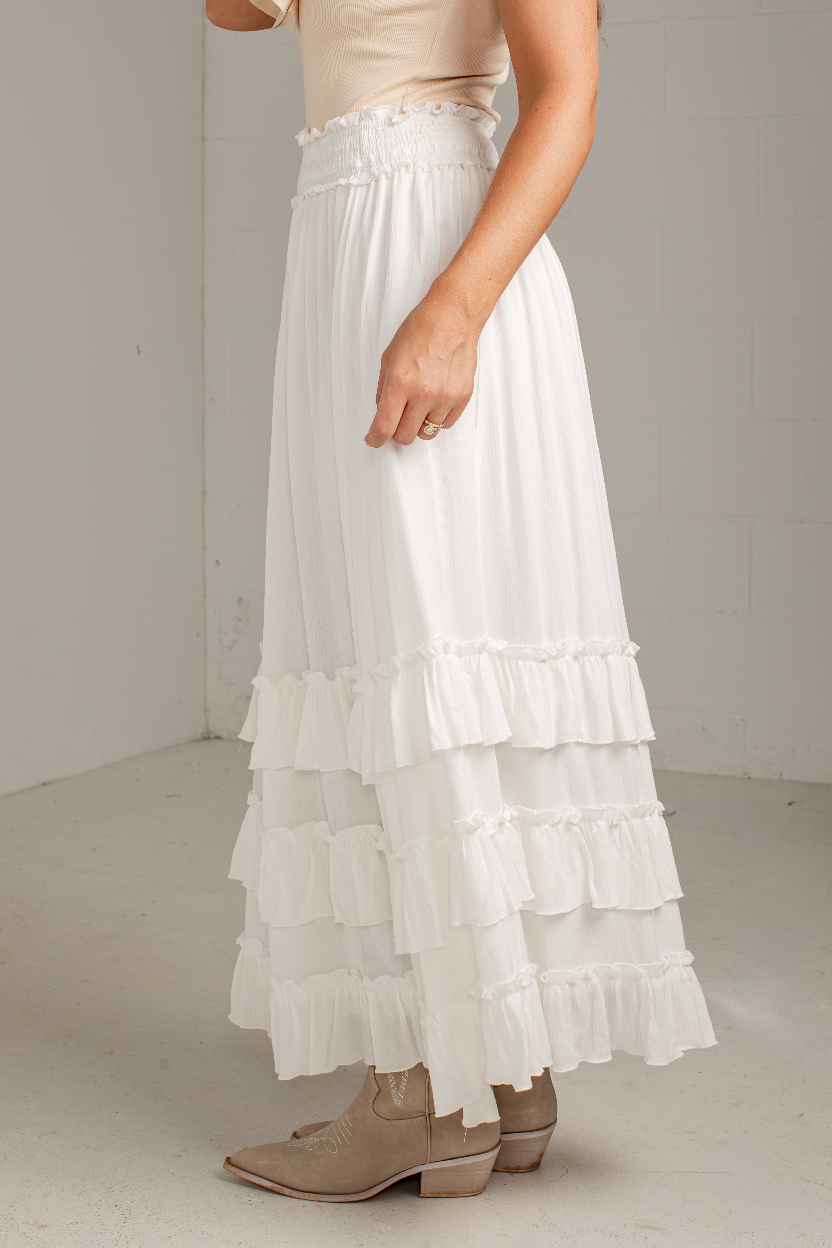 A white tiered maxi skirt with a ruffled waistband, worn by a model with beige boots and a beige top in a neutral studio setting.