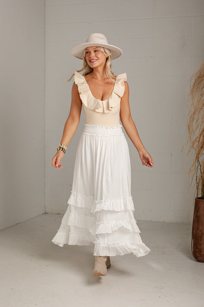 A woman wearing a cream ruffled sleeveless top and a long white tiered skirt, paired with a wide-brimmed cream hat and beige boots, standing in a bright studio with a white wall.