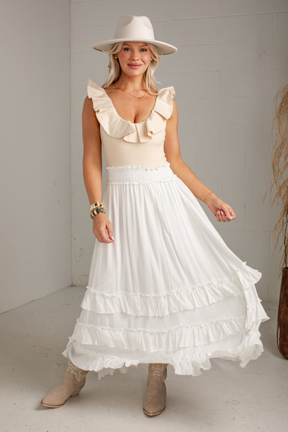 A woman modeling a bohemian-style white tiered maxi skirt with ruffle details, paired with a cream ruffled top, beige hat, and beige boots, standing in a minimalist white room.