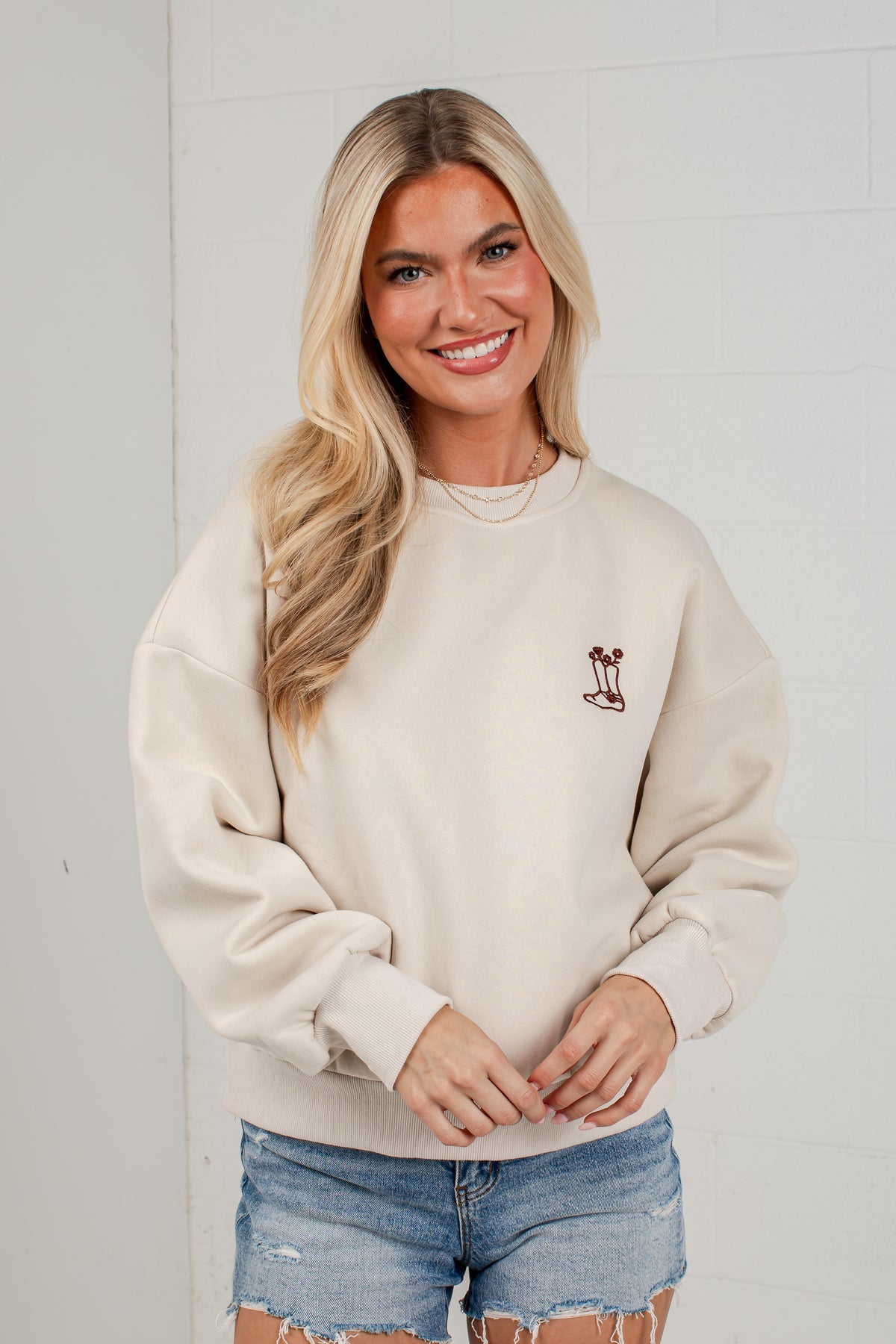 A smiling blonde woman wearing a cream-colored crewneck sweatshirt with a small embroidered palm tree graphic on the left chest, paired with distressed denim shorts, standing against a white brick wall.