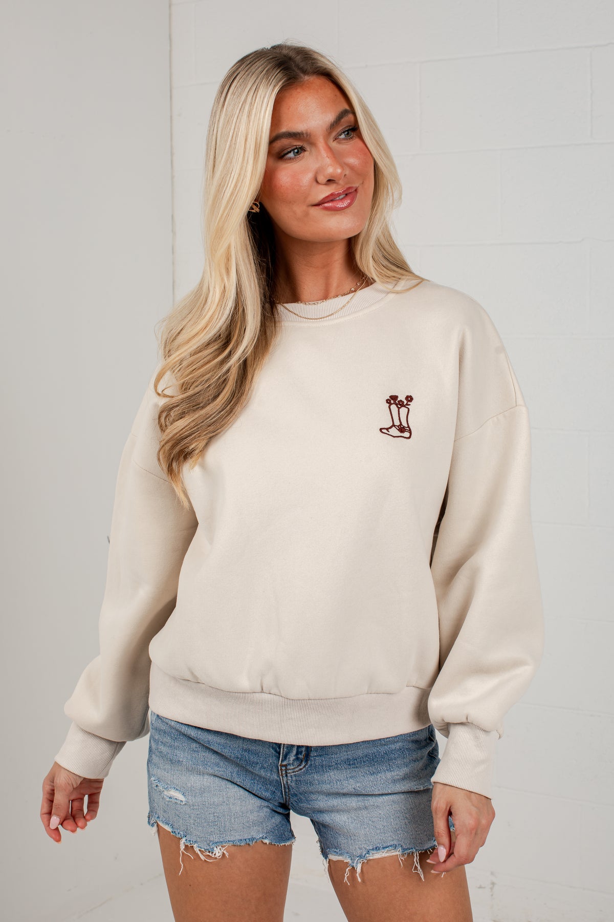 A blonde woman wearing a cream-colored crewneck sweatshirt with a small embroidered graphic on the left chest, paired with distressed denim shorts, standing against a white brick wall.