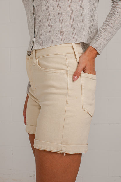A person wearing light beige rolled denim shorts with a gray ribbed knit top, standing against a white brick wall.