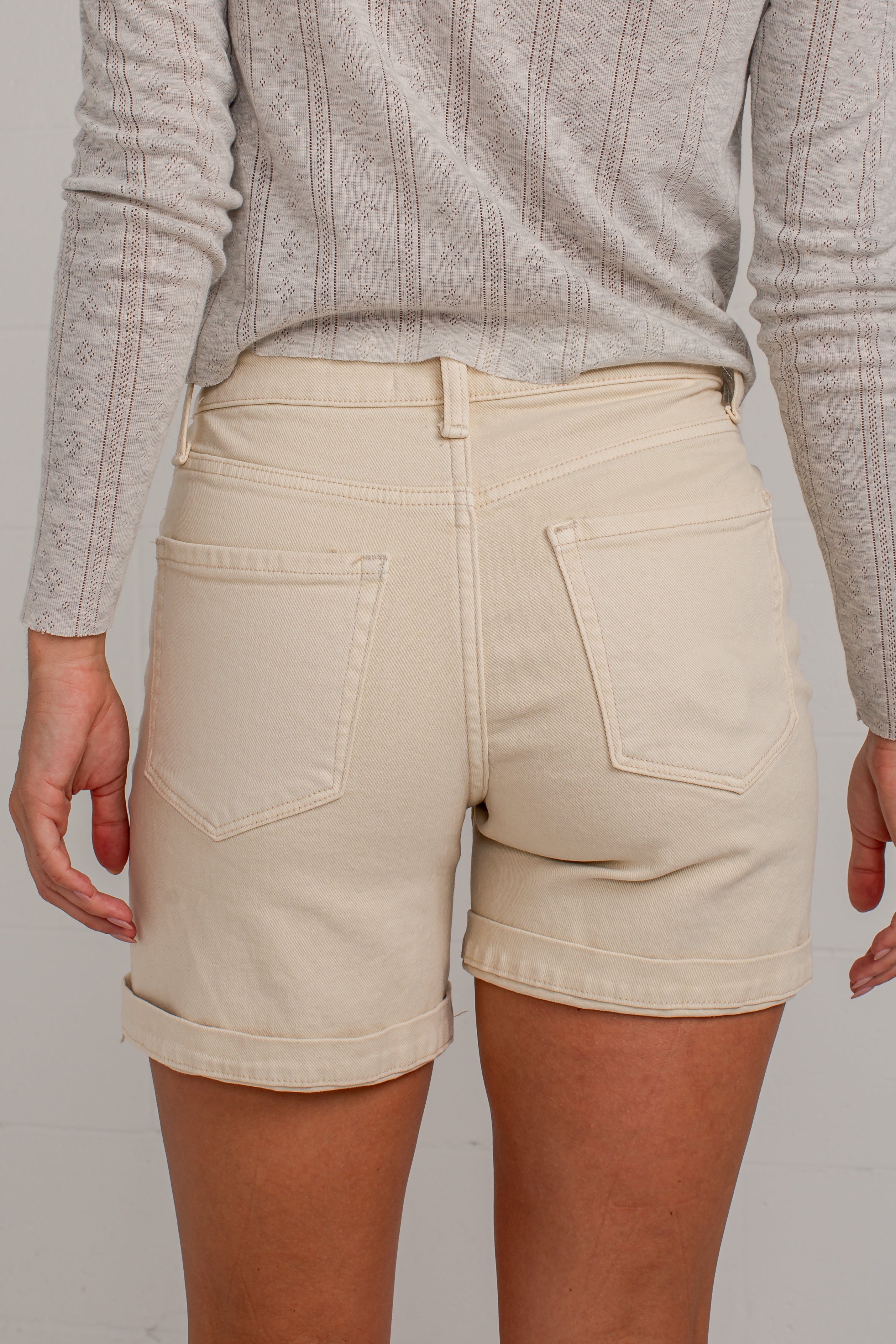 A woman modeling a light gray long-sleeve button-up shirt and cream-colored denim shorts, standing against a white studio wall.