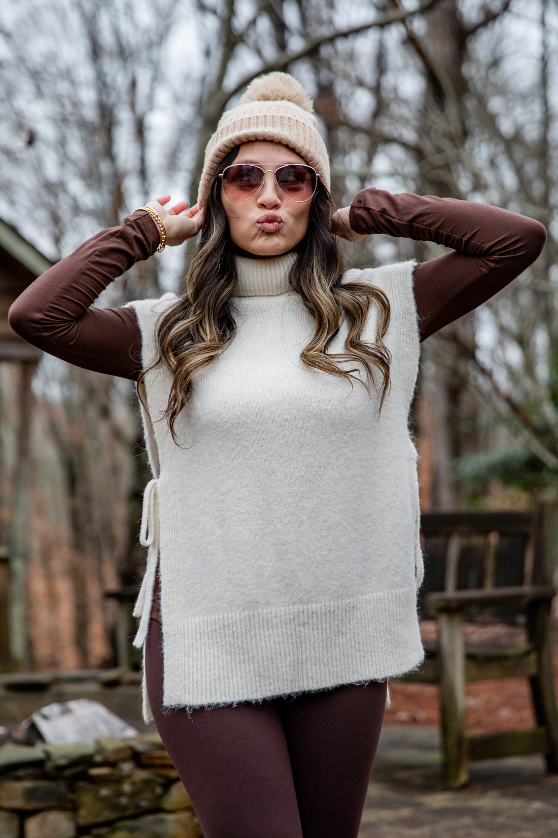 Woman wearing a white sleeveless sweater over a brown long-sleeve shirt, sunglasses, and a beige beanie outdoors.