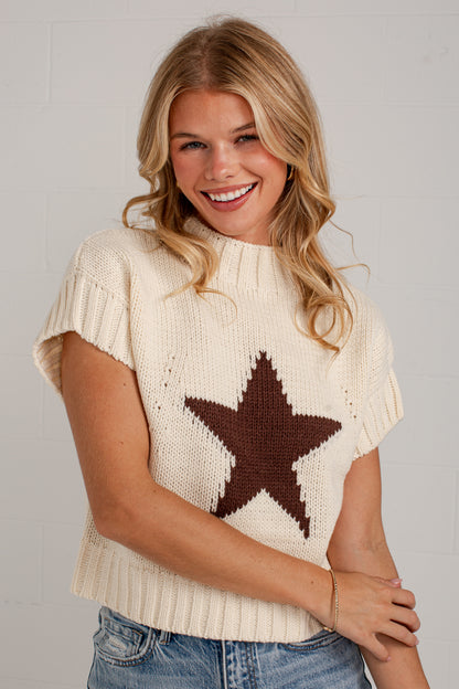 A smiling blonde woman wearing a cream knit sweater with a large dark brown star on the front, paired with blue jeans, standing against a light gray brick wall.