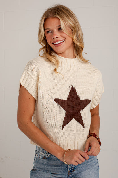 A woman wearing a cream-colored knit sweater with a large brown star pattern on the front, paired with blue jeans, standing against a white brick wall.