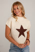 A woman wearing a cream-colored knit sweater with a large brown star pattern on the front, paired with blue jeans, standing against a white brick wall.