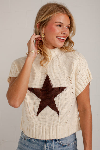 A woman wearing a cream-colored knit sweater with a large dark brown star on the front, paired with blue jeans, standing against a white brick wall.