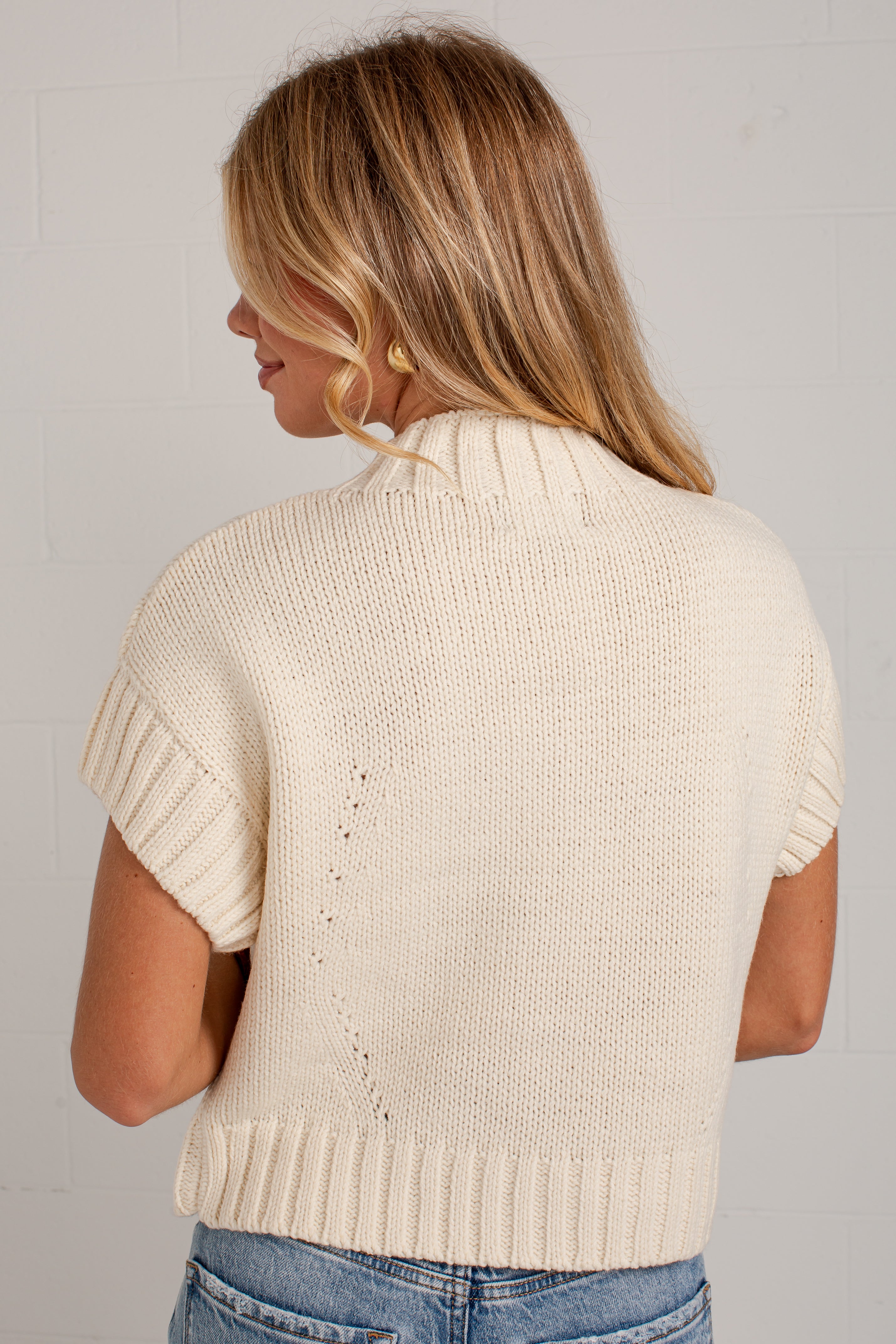 A woman viewed from the back wearing a cream-colored short-sleeve knit sweater with ribbed trim, paired with blue jeans, against a light gray brick wall.