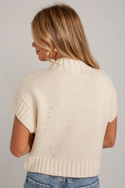 A woman viewed from the back wearing a cream-colored short-sleeve knit sweater with ribbed trim, paired with blue jeans, against a light gray brick wall.