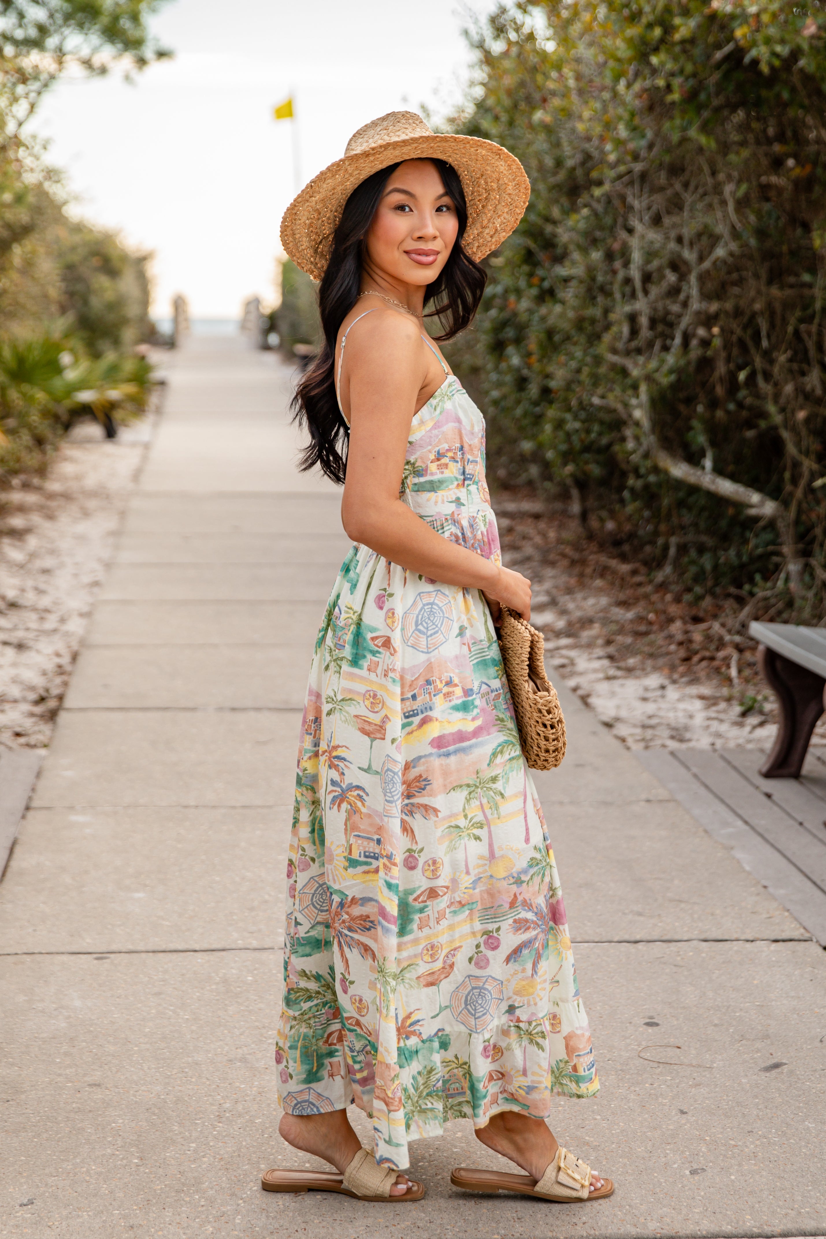 Woman in a floral dress and hat walking on a path with greenery.