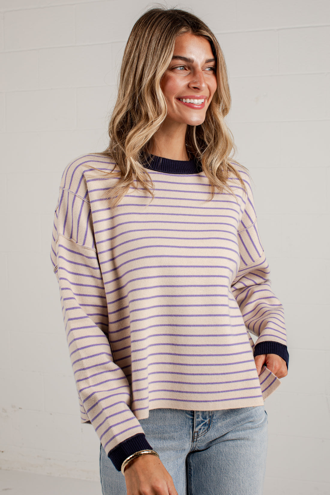 A woman wearing a long-sleeve knit top with thin horizontal stripes in beige and purple, featuring dark navy ribbed trim at the crew neck, cuffs, and hem, paired with light-wash blue jeans.