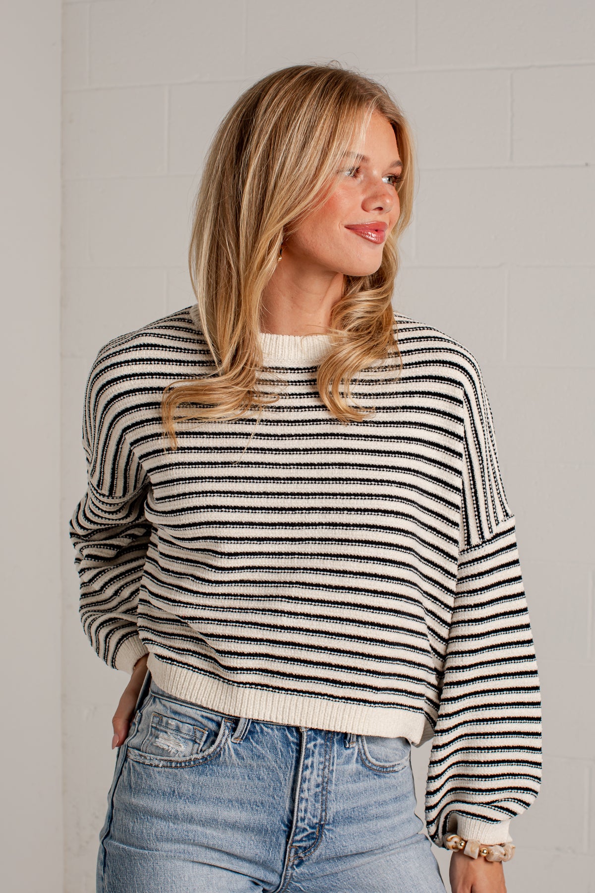 A woman wearing a black and off-white horizontally striped knit sweater with balloon sleeves, paired with light-wash blue jeans, standing against a light gray brick wall.
